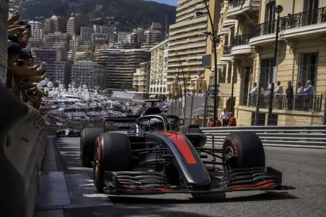 Monoposto di Formula 1 in una stretta curva cittadina con barriere, palazzi e porto pieno di yacht sullo sfondo